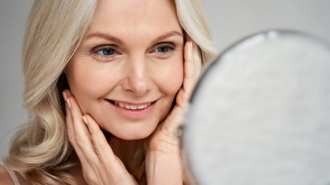 Smiling woman examining her facial skin in a mirror while gently touching her cheeks.